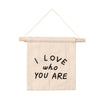The I Love Who You Are Canvas Sign - Natural is a beige fabric banner on a wooden rod with "I LOVE who YOU ARE" in black, offering inspirational Kenyan artisan decor and adding warmth to any space.