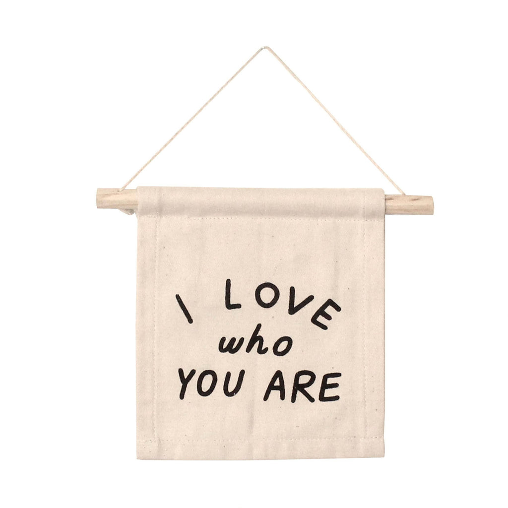 The I Love Who You Are Canvas Sign - Natural is a beige fabric banner on a wooden rod with "I LOVE who YOU ARE" in black, offering inspirational Kenyan artisan decor and adding warmth to any space.