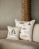 The light beige sofa is adorned with the Coral & Tusk Sailing Foxes Pillow, featuring playful boat and fox illustrations, alongside another beige throw pillow. A tall, textured decorative object appears blurred in the background.