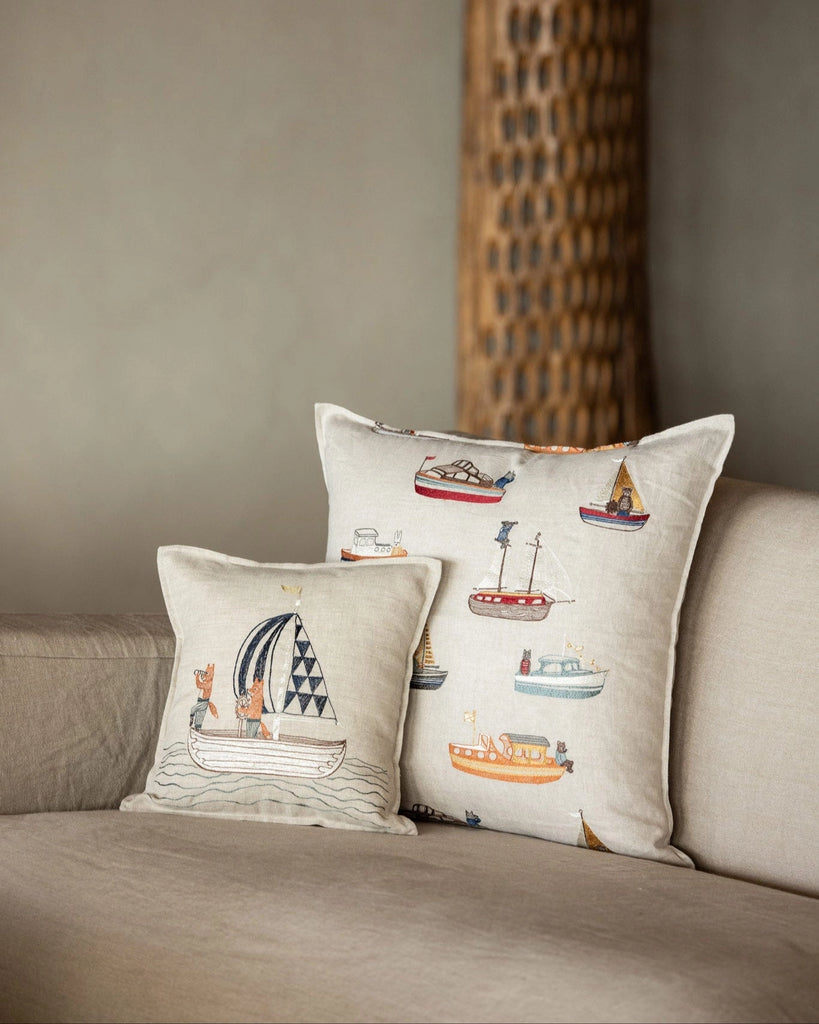 The light beige sofa is adorned with the Coral & Tusk Sailing Foxes Pillow, featuring playful boat and fox illustrations, alongside another beige throw pillow. A tall, textured decorative object appears blurred in the background.