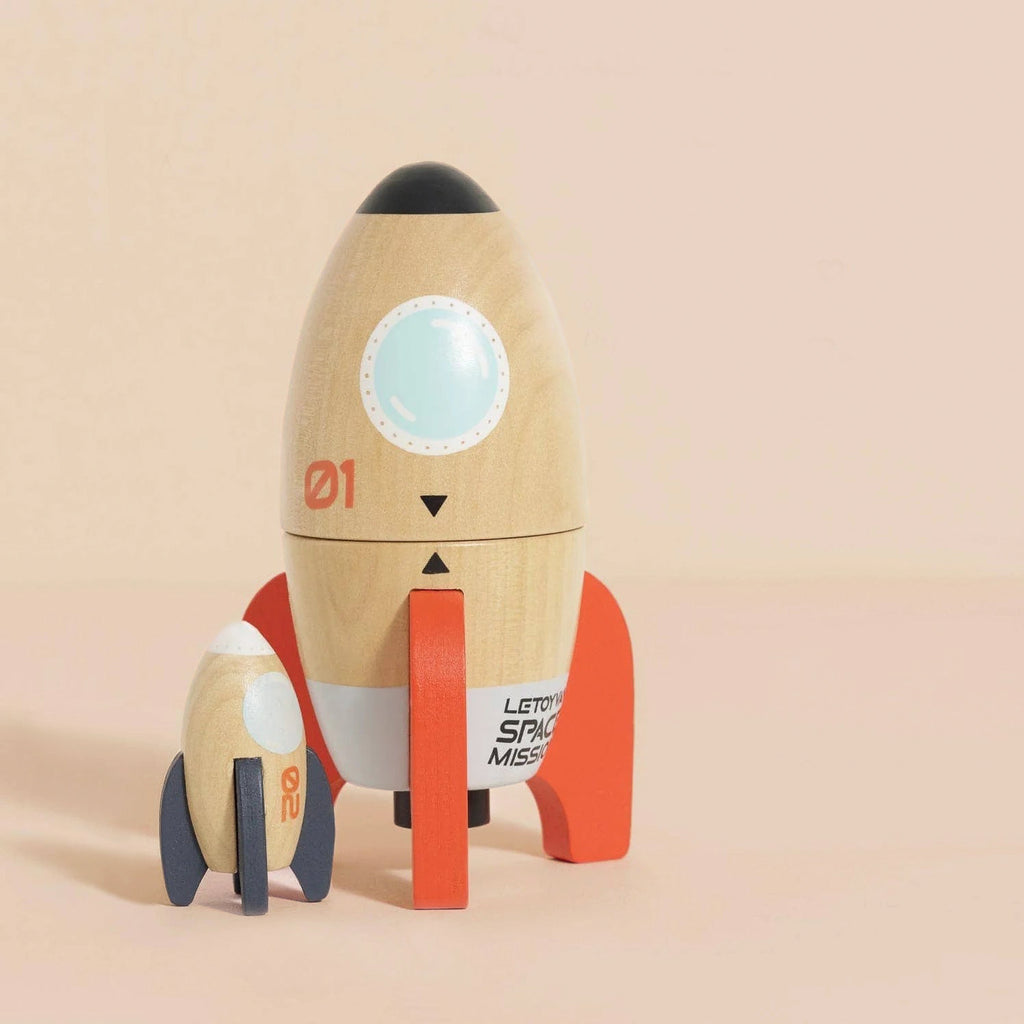 The Magnetic Space Rocket Duo features two wooden toy rockets with red fins and window details, standing upright on a light pink surface. Their playful, minimalist design is ideal for fans of plastic-free toys. Ships in one week.