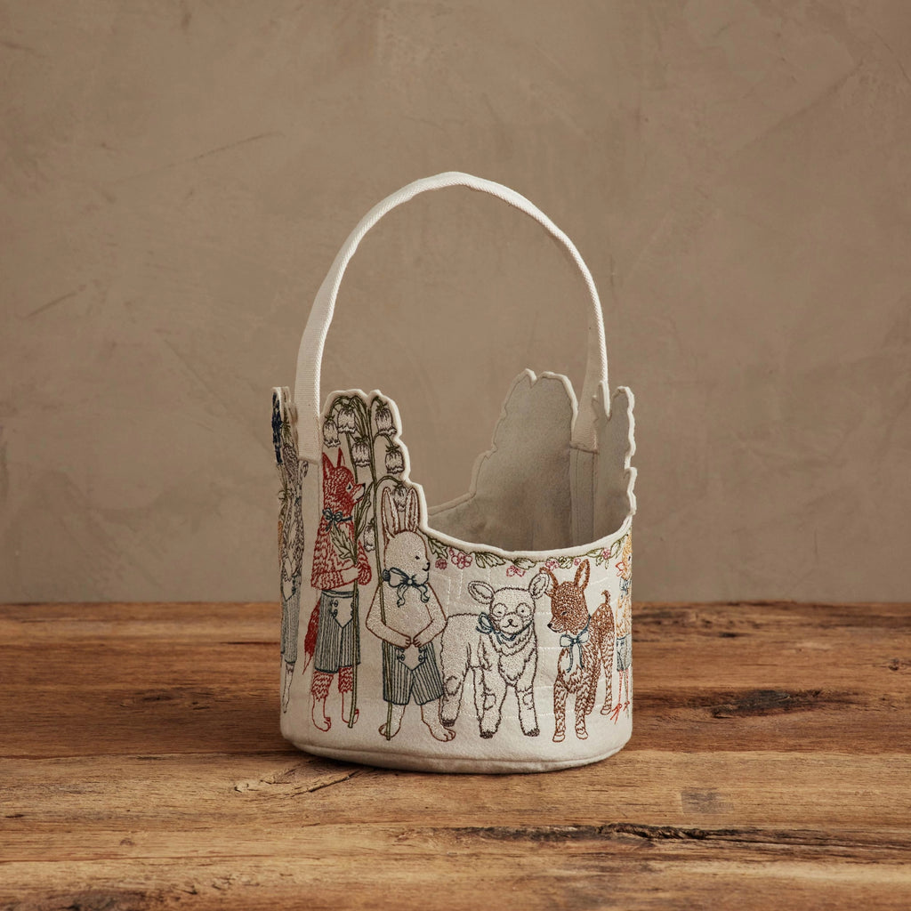 The Coral & Tusk Easter Parade Basket, featuring a scalloped edge, handle, and embroidered animals and children, rests on a rustic wooden surface against a neutral textured background.