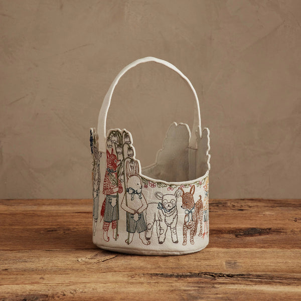 The Coral & Tusk Easter Parade Basket, featuring a scalloped edge, handle, and embroidered animals and children, rests on a rustic wooden surface against a neutral textured background.
