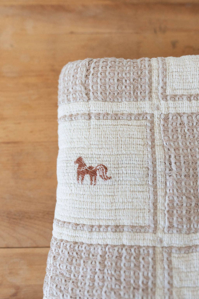 A folded, textured beige Organic Cotton Twin Size Blanket - Pony, featuring a delicate crochet trim and a small brown embroidered pony on one section, is placed on a wooden surface.