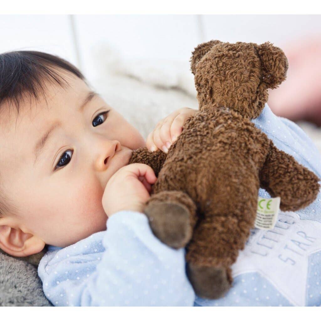A baby in a blue outfit lies on a soft surface, holding and chewing on the Senger Naturwelt Stuffed Animal - Brown Bear, gazing off to the side.