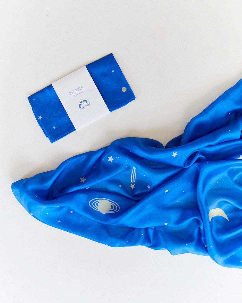 A folded blue pure silk scarf with a white label sits beside the Sarah's Silks Enchanted Playsilk - Starry Night, unfurled and decorated with planets, stars, and a crescent moon—perfect for open-ended play against a white background.
