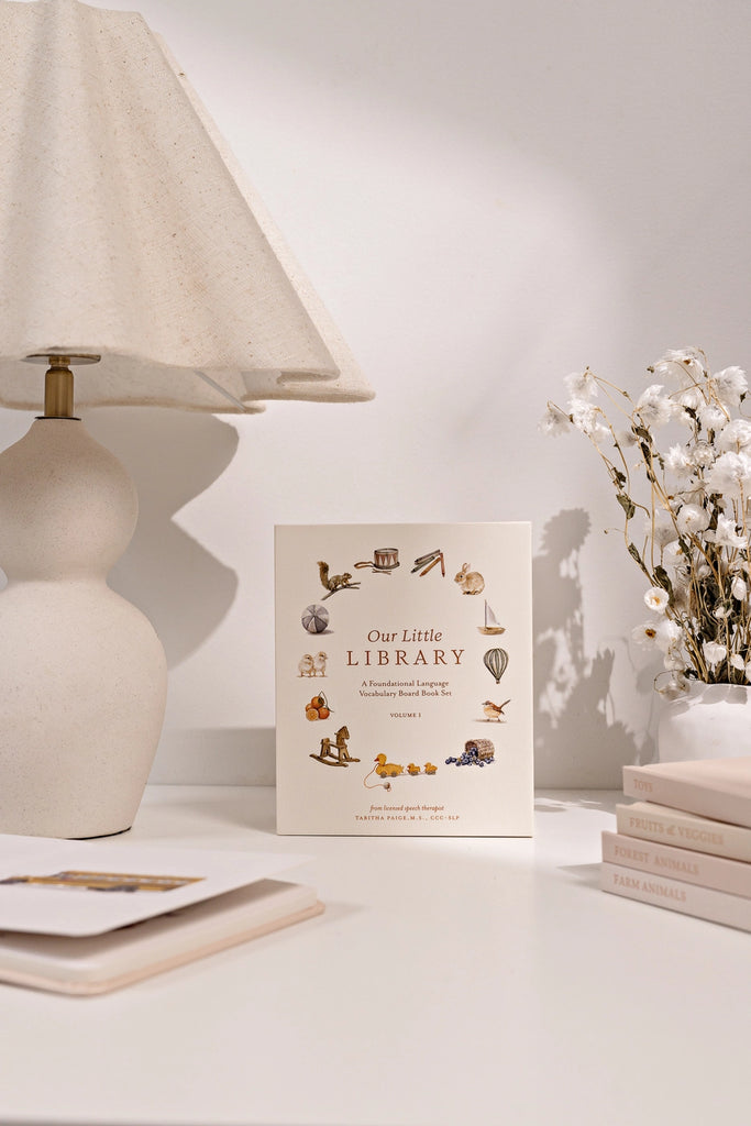 A cozy reading nook with a decorative lamp on the left and a vase of cotton branches on the right. Between them stands an illustrated card titled "Our Little Library Book Box Set" surrounded by a few small board