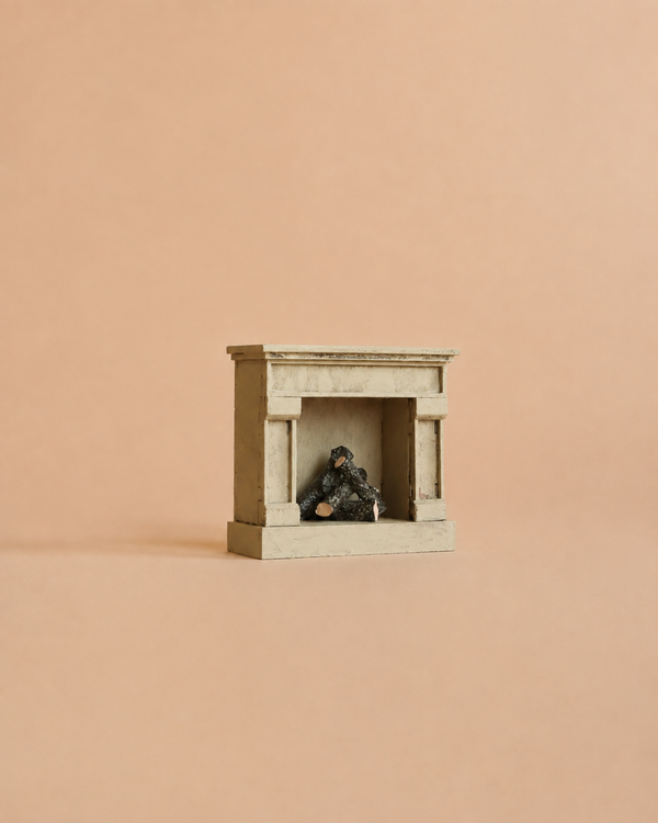 The Maileg Fireplace, Mouse Size - Off White is centered against a plain peach background, its small white wooden frame with a vintage finish and miniature logs inside evoking the charm of a cozy miniature home.