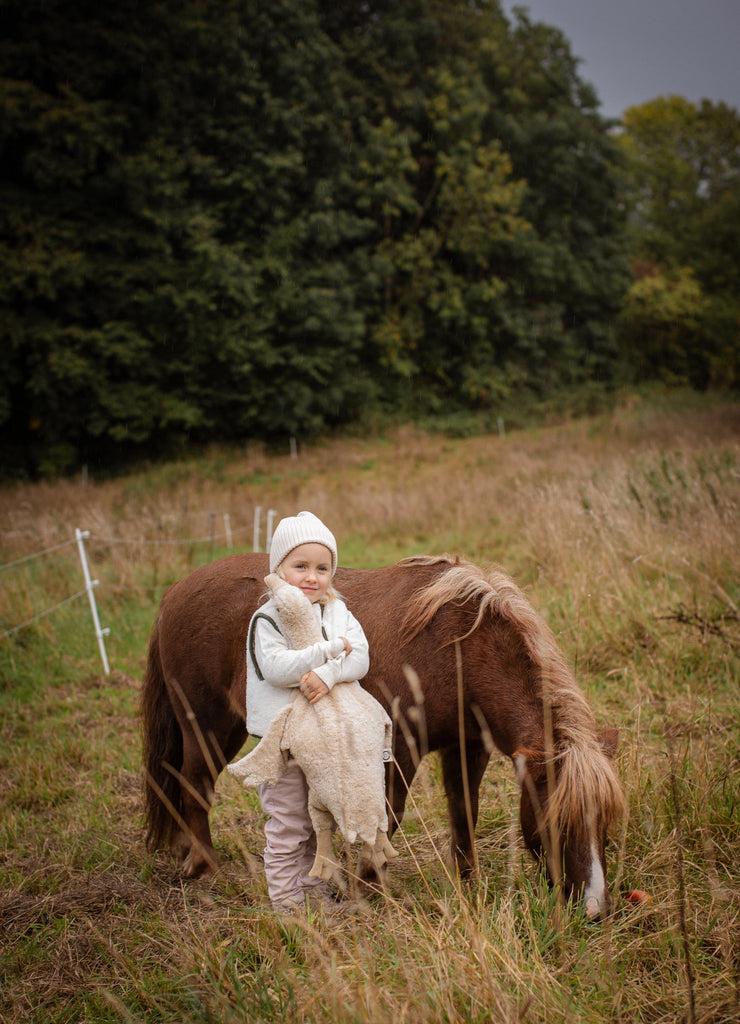 A small child in a white hat and cozy GOTS-certified cotton clothes stands in a grassy field beside a brown and white pony, holding the Senger Naturwelt Cuddly Animal - Linen Goose, with dense green trees in the background.