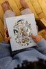 A person in a blue sweater holds the "Midnight Mail 500 Piece Puzzle" box, which features a whimsical winter scene with animals, trees, and snow. Their bare feet are visible on the wooden floor.