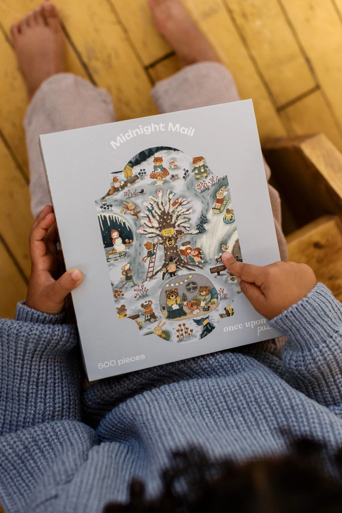 A person in a blue sweater holds the "Midnight Mail 500 Piece Puzzle" box, which features a whimsical winter scene with animals, trees, and snow. Their bare feet are visible on the wooden floor.