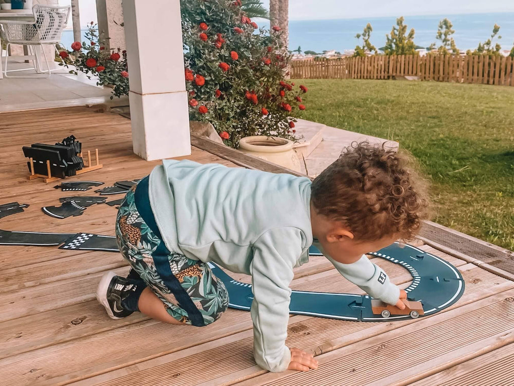 A young child in a light blue shirt and patterned shorts plays with the Large Flexible Toy Race Track - Grand Prix and a toy car on a wooden deck outside, with flowers and a garden visible in the background.