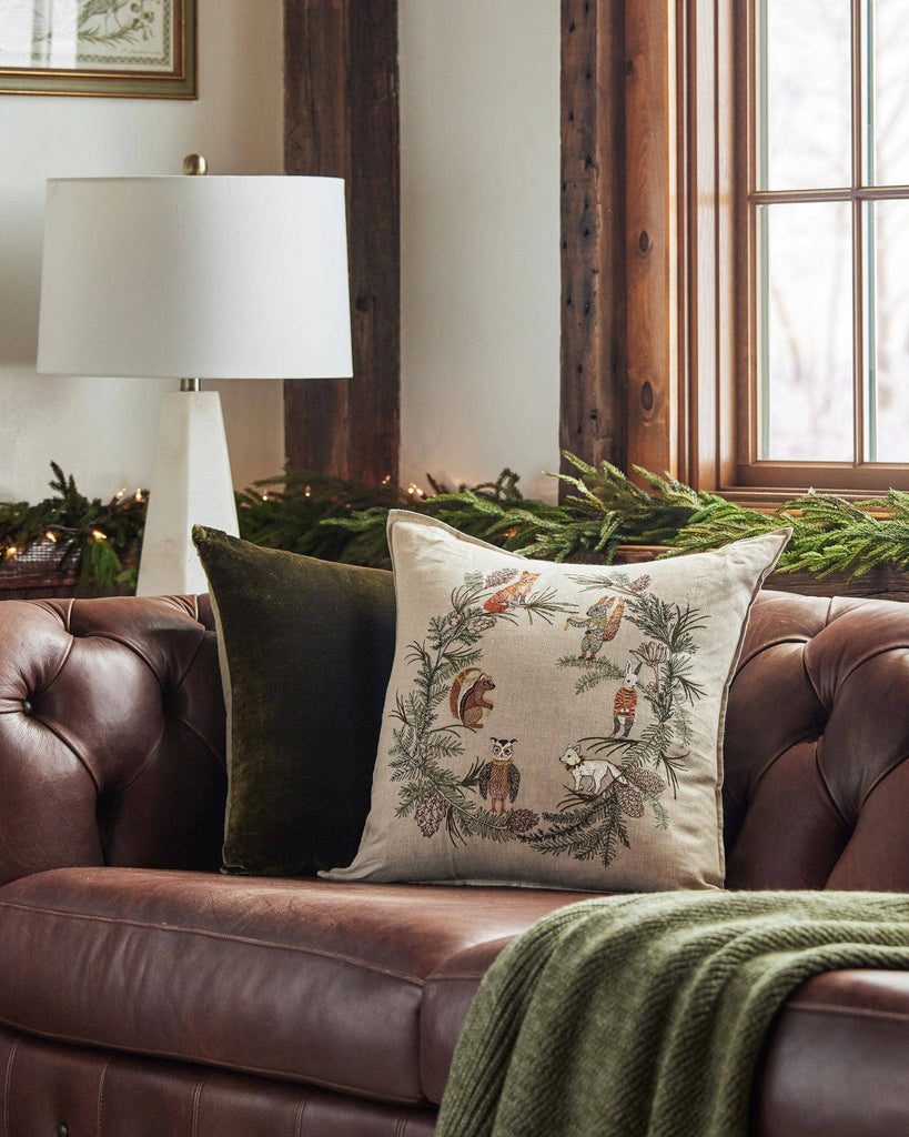 A brown leather sofa with a green throw and two cushions—one plain green and one Coral & Tusk Tree Trimmers Wreath Pillow—stands by a window and white lamp, with pine garland festooning the festive windowsill.