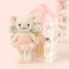 Cuddle + Kind Tiny Lily the Fairy, a handmade fair trade doll with wings in a pink dress and gold star wand, stands before a pastel floral fairy house on a soft pink background.