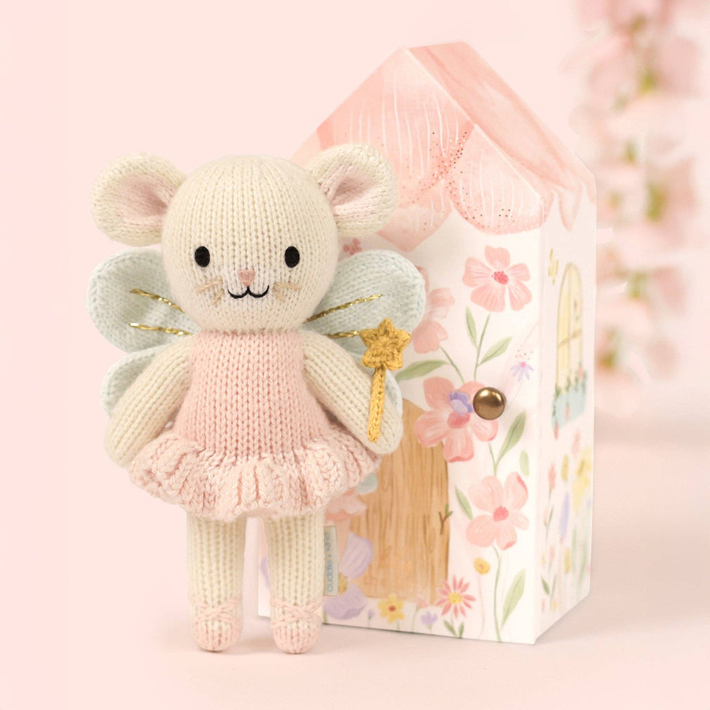 Cuddle + Kind Tiny Lily the Fairy, a handmade fair trade doll with wings in a pink dress and gold star wand, stands before a pastel floral fairy house on a soft pink background.