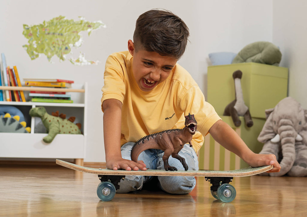 A young boy in a yellow shirt kneels on the floor, excitedly playing with the Schleich Giganotosaurus Large Dinosaur Toy with Movable Jaw, which is perched on a skateboard in a child’s room decorated with stuffed animals and books.