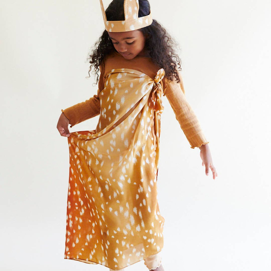 A young girl with long curly hair wears Sarah's Silks Animal Print Playsilk - Fawn, featuring brown fabric with white spots and a matching headband with deer-like ears, as she looks down and holds out part of her dress.