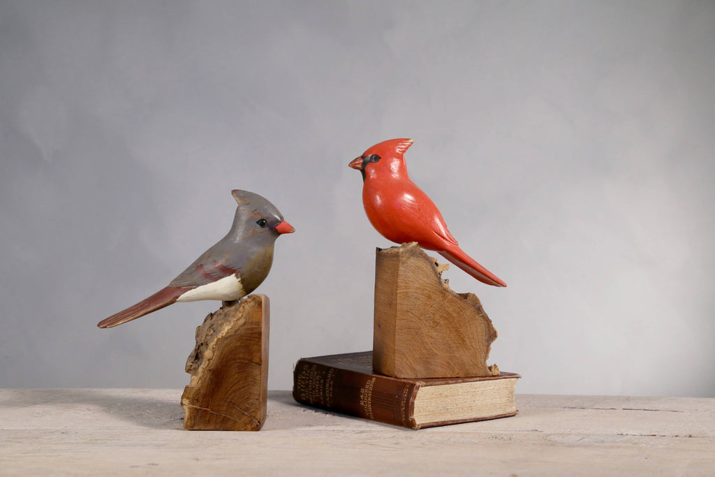 The Hand Carved Ornament - Cardinal Bird-Female (7"H) perches on a rustic wood stand atop an old book, creating a charming display against a gray background—perfect as an anniversary gift.