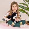 A young girl with long brown hair sits on a pink mat, holding a cuddly Teddy Hermann Black Cat Stuffed Animal. She is wearing a pink sweater with animal prints, green pants with heart patches, and striped socks. Behind her is a large green leafy plant and a woven basket.