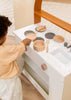 A child plays with the Wooden Pots and Pans Playset (6 Pcs), reaching for a pot on the stovetop. Other wooden cooking toys and cookware are neatly arranged on the counter, creating a delightful scene with this charming playset.