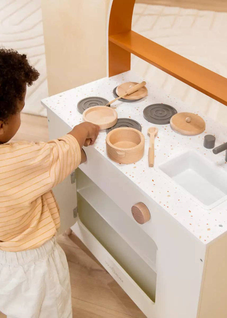A child plays with the Wooden Pots and Pans Playset (6 Pcs), reaching for a pot on the stovetop. Other wooden cooking toys and cookware are neatly arranged on the counter, creating a delightful scene with this charming playset.
