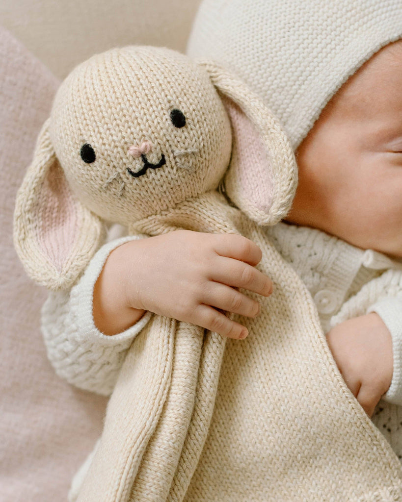 A baby dressed in cream cuddles the Cuddle + Kind Organic Cotton Baby Bunny Lovey (Oatmeal), featuring embroidered eyes, nose, and whiskers—a soft, fair trade toy that makes an adorable gift.