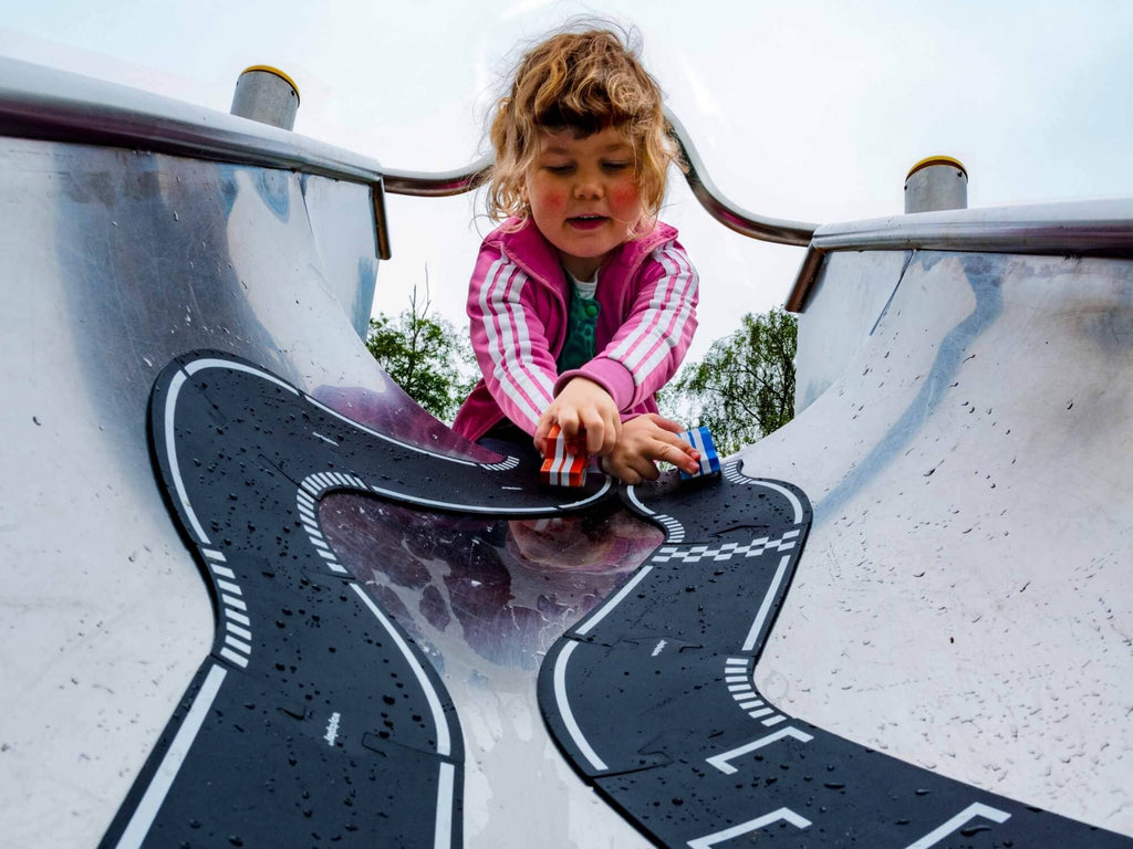 A young child in a pink jacket plays with cars on the Large Flexible Toy Race Track - Grand Prix, designed to look like a racetrack, as seen from the bottom of the slide looking up.