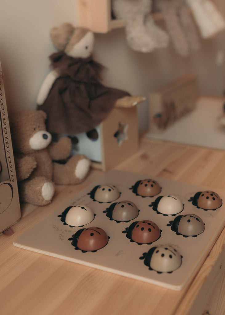 The My Little Lady Bug Wooden Puzzle, featuring ladybug-shaped pieces, sits on a wooden table surrounded by plush toys—including a teddy bear and a doll in a brown dress—in a cozy, softly lit children’s room, offering a charming example of Montessori-inspired toys that encourage hands-on learning and play.