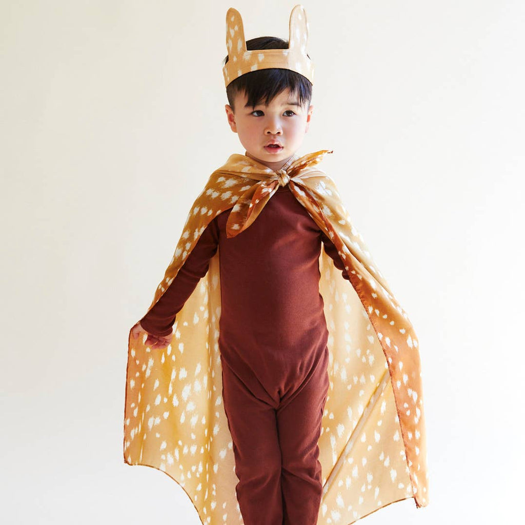 A young child wears a brown bodysuit, cape, and deer ear headband, resembling a fawn while standing against a white background—perfect for imaginative play with Sarah's Silks Animal Print Playsilk - Fawn.