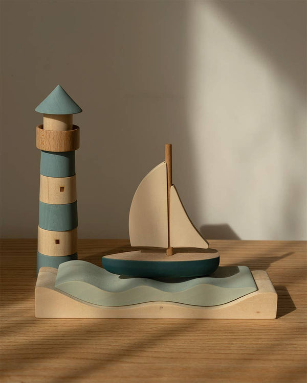 The Sabo Concept Sea Adventure Set features a blue and beige striped lighthouse, a white-sailed sailboat, and wavy water on wood, evoking a maritime playtime in gentle natural light.
