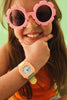 A smiling child with long brown hair and a bright orange top rests their chin on their hand, wearing pink flower-shaped sunglasses and the Mini Kyomo Watch - Watercolor. Green background.