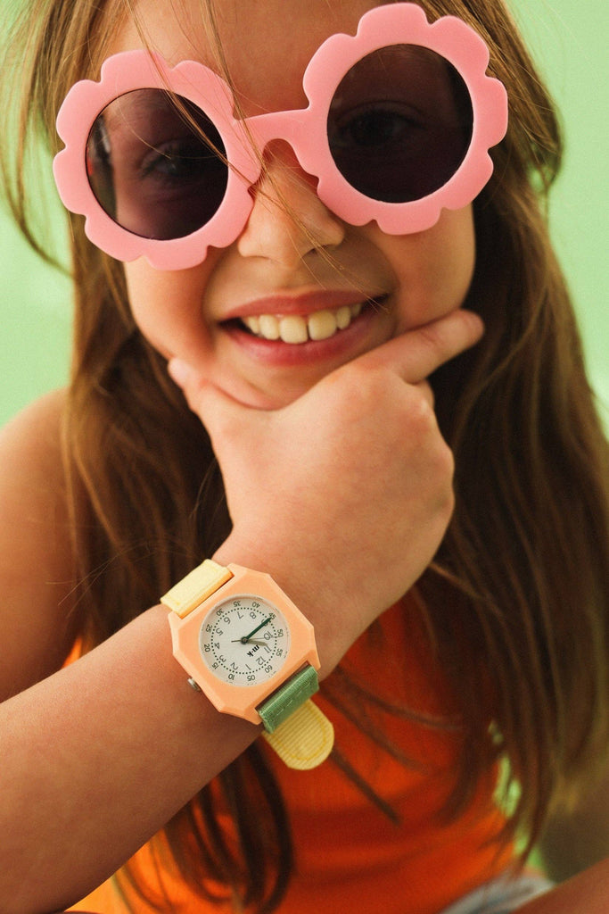 A smiling child with long brown hair and a bright orange top rests their chin on their hand, wearing pink flower-shaped sunglasses and the Mini Kyomo Watch - Watercolor. Green background.