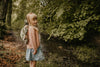 A smiling blonde girl stands by a forest creek in a pink sweater, blue skirt, and the Children's Backpack - Turtle, featuring adjustable shoulder straps. She is surrounded by lush green trees and foliage.