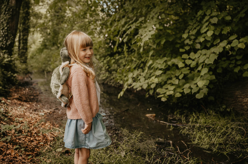 A smiling blonde girl stands by a forest creek in a pink sweater, blue skirt, and the Children's Backpack - Turtle, featuring adjustable shoulder straps. She is surrounded by lush green trees and foliage.