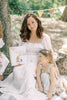 A woman in a white dress holds Winnie-the-Pooh: Finding Feelings as a young girl in stripes leans on her. They sit by a tree outdoors, surrounded by sunlight and bunting—perfect for capturing heartfelt moments with toddlers.