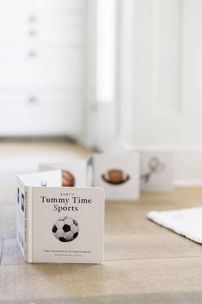 The "Baby's Tummy Time Sports" high-contrast board book, featuring a soccer ball on the cover, stands open on a wooden floor for visual stimulation; two more open baby books appear blurred in the background.