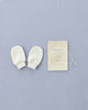 A pair of Baby Mitten organic cotton baby mittens designed to prevent babies from scratching are placed next to a small beige drawstring pouch with text that reads "Baby Mitten," “100% Cotton,” and a heart illustration. They are set against a light blue fabric background, perfect for infants aged 0-3 months.