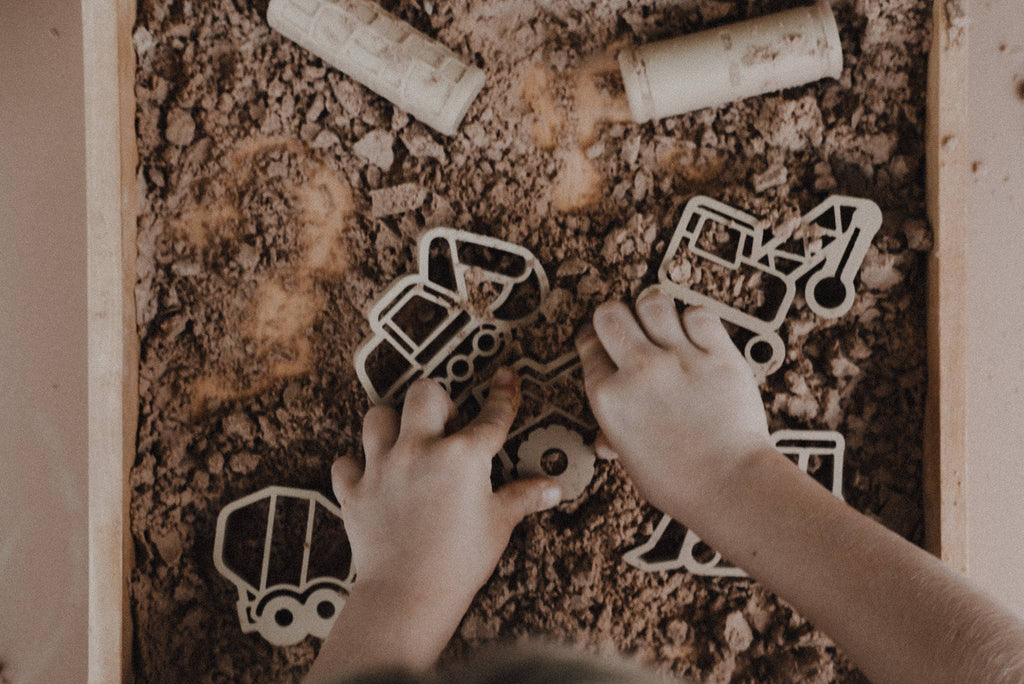 A child’s hands play with the Construction Cutter Set - Eco Playdough Tool in a tray filled with brown dirt, using the playdough tools to shape a construction-themed scene. Several other vehicle cutters and rolling pins from the set are also visible on the dirt surface, making this sensory play experience engaging and creative.