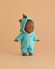 The Minikane Mini Pancha Doll - Arsène with Dinosaur Jumpsuit features a high-quality vinyl doll with brown skin, dressed in a fuzzy teal onesie adorned with playful dinosaur spikes on the hood, posed against a plain beige background—perfect for inspiring imaginative play.