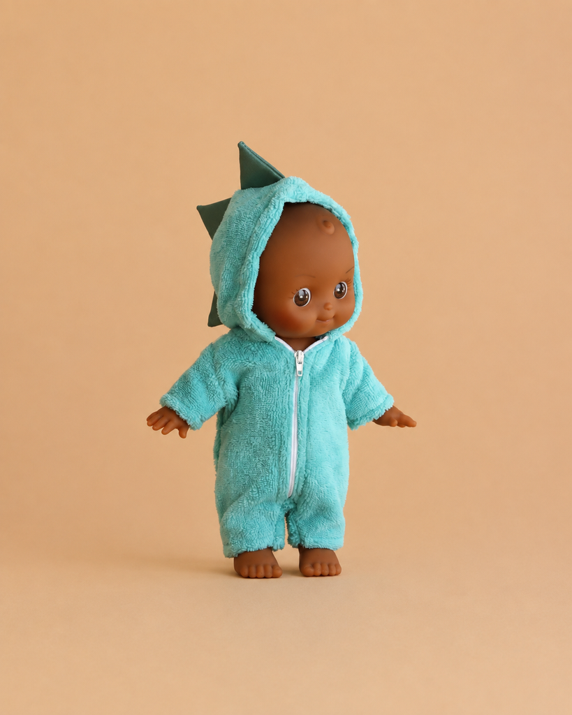The Minikane Mini Pancha Doll - Arsène with Dinosaur Jumpsuit features a high-quality vinyl doll with brown skin, dressed in a fuzzy teal onesie adorned with playful dinosaur spikes on the hood, posed against a plain beige background—perfect for inspiring imaginative play.