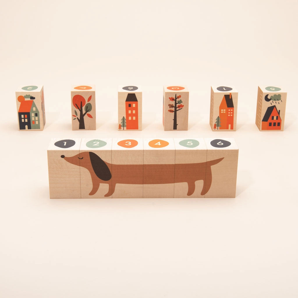 A set of Uncle Goose Environments Neighborhood Blocks, featuring a cute dachshund divided into sections numbered 1 to 6, positioned in front of blocks illustrated with colorful trees and houses on a white background.