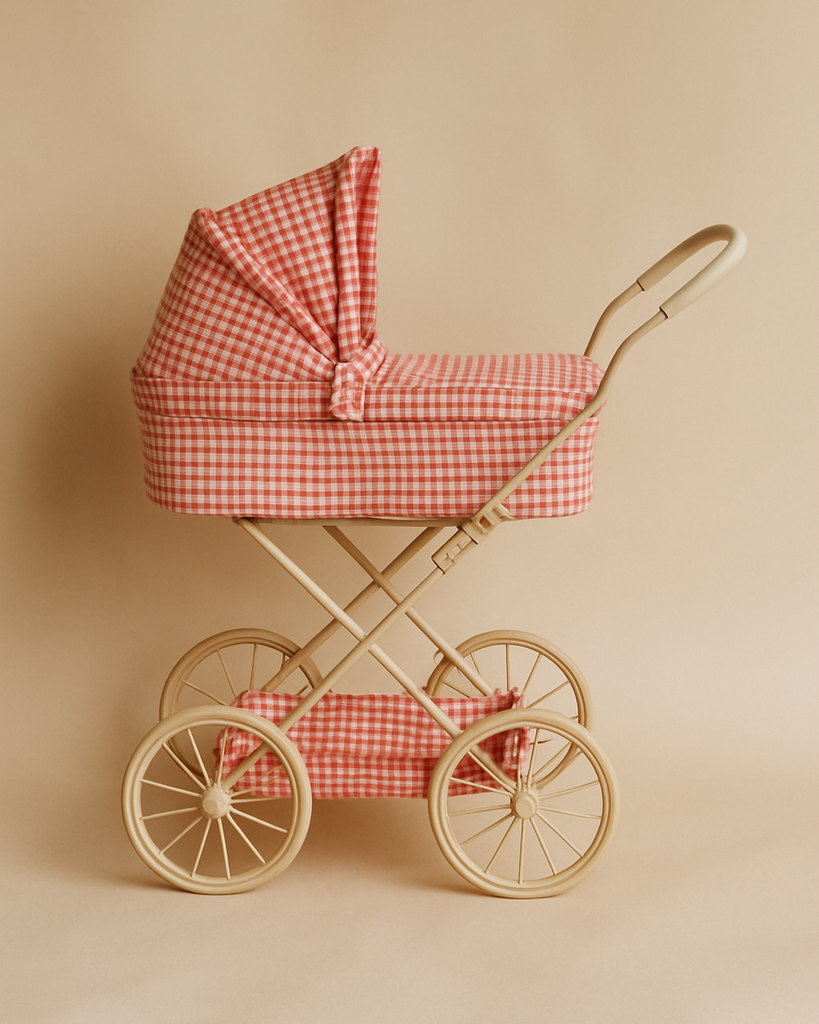 The Konges Sloejd Doll Pram - Bubblegum Check features red and white quilted fabric, a beige frame, four large wheels, a matching storage basket, and a padded mattress, all set against a beige background.