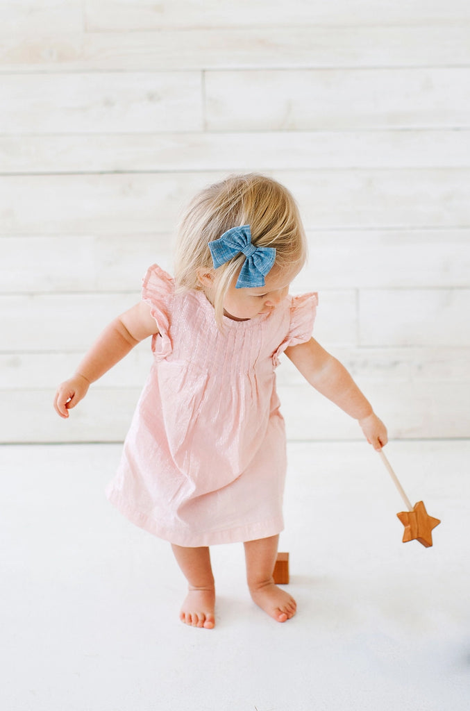 A toddler girl in a pink dress and blue bow, tentatively walking and holding a Bannor Toys Wooden Star Wand, against a white wooden background.