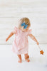 A toddler girl in a pink dress and blue bow, tentatively walking and holding a Bannor Toys Wooden Star Wand, against a white wooden background.