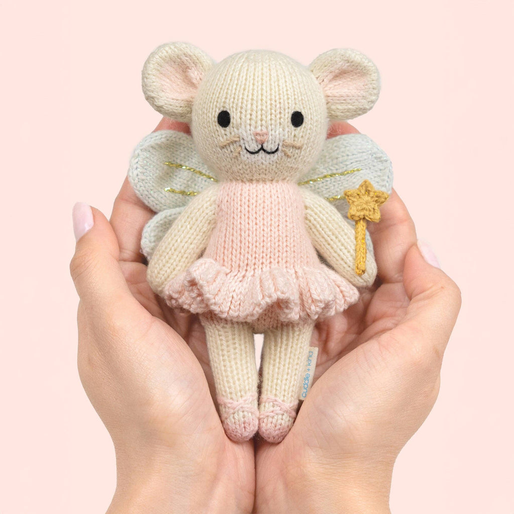 Two hands gently hold a Cuddle + Kind Tiny Lily the Fairy doll, wearing a pink dress and fairy wings while holding a yellow star wand, against a pale pink background.