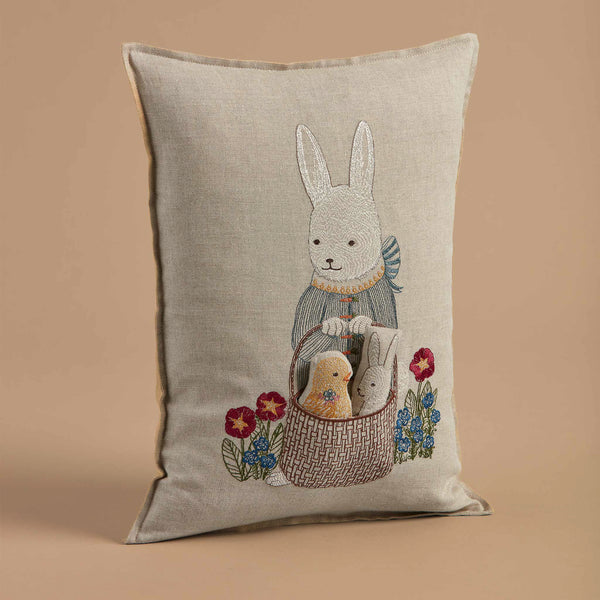 The Coral & Tusk Easter Bunny Pocket Pillow features an embroidered Easter Bunny with a basket and two smaller rabbits, set against a light brown background with colorful flowers, capturing a cherished Easter tradition.