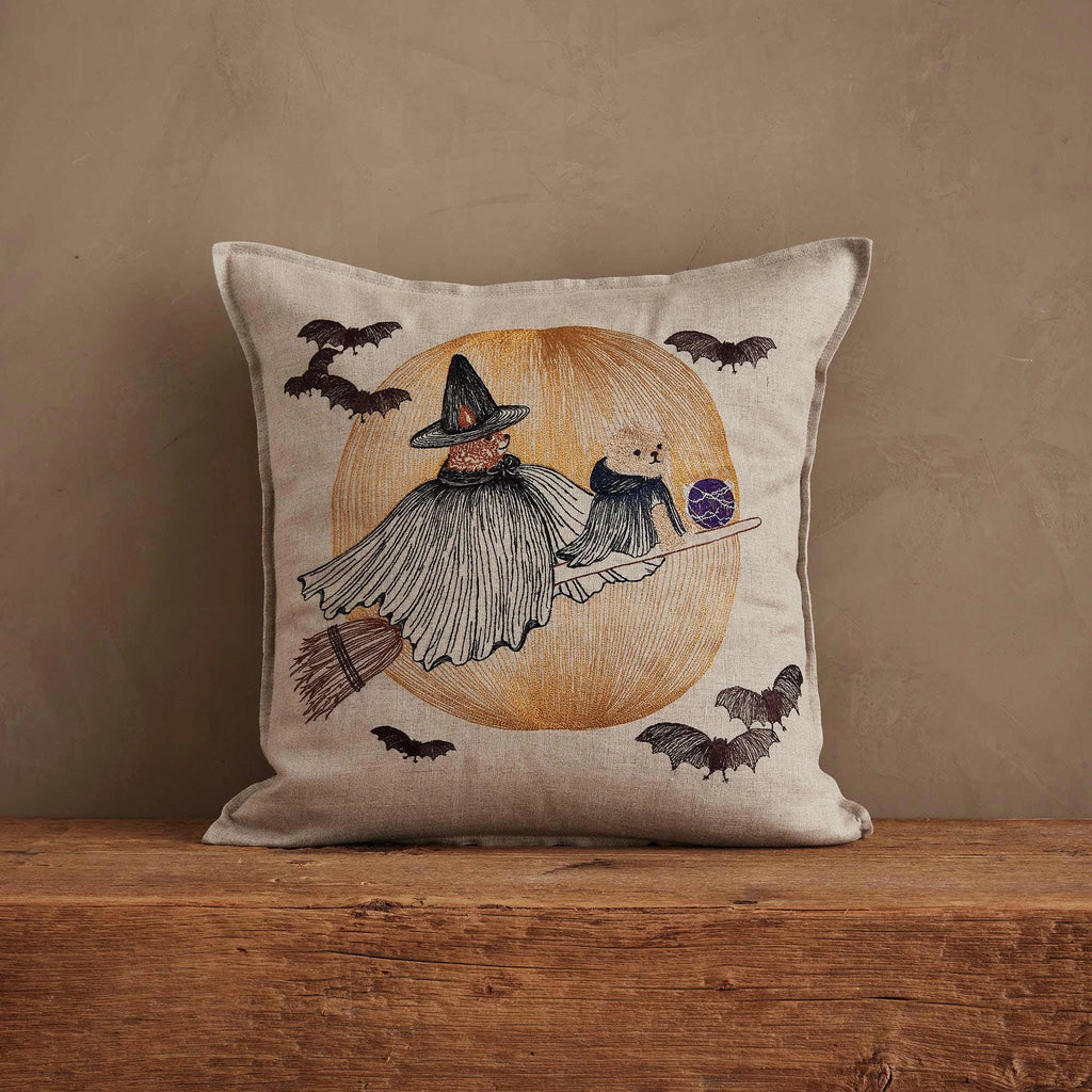 The Coral & Tusk Supermoon Spell Pillow features a witch and cat riding a broomstick before a large orange moon with bats, adding enchanting charm to your space against a plain brown background.