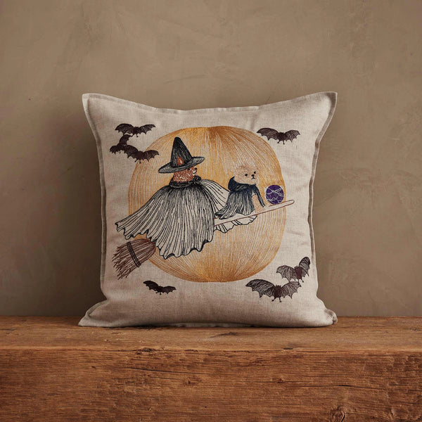 The Coral & Tusk Supermoon Spell Pillow features a witch and cat riding a broomstick before a large orange moon with bats, adding enchanting charm to your space against a plain brown background.