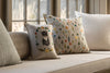 Three decorative pillows rest on a beige window seat: the Coral & Tusk Kitten Cameo Pillow featuring an embroidered cat, a floral-patterned pillow ideal for spring decor, and a plain white one. Sunlight pours in from the window behind them.