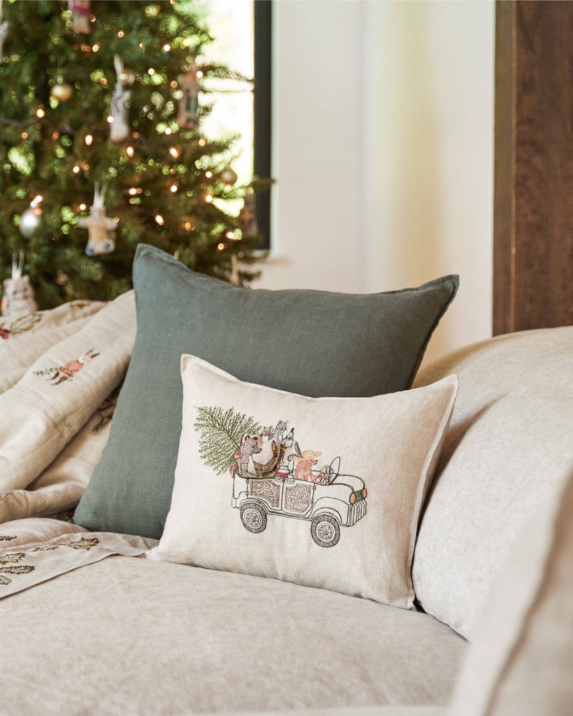 A cozy beige sofa holds the Coral & Tusk Christmas Tree Car Pocket Pillow, featuring woodland animals driving a truck with a Christmas tree, while a beautifully lit Christmas tree adds festive flair in the background.